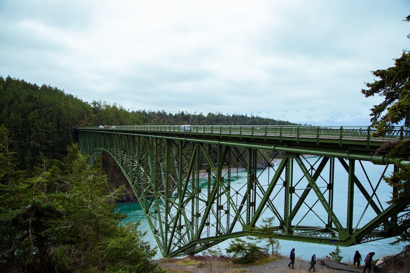 Green steel bridge spans forested canyon above clear blue river water